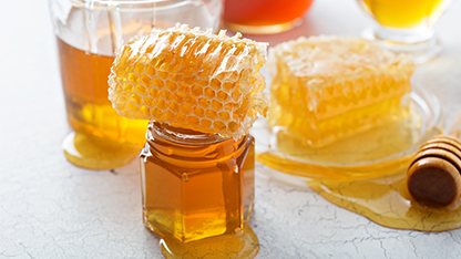 A small glass jar of honey filled with clear yellow honey with a square segment of honeycomb placed on top of the jar