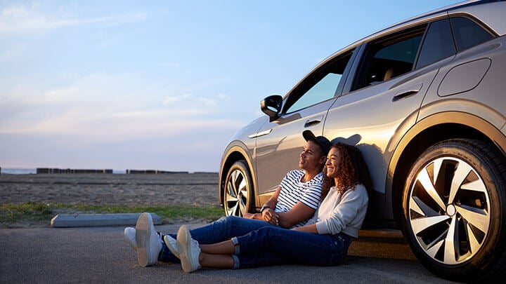 A young man and woman sit holding hands against a car by the beach