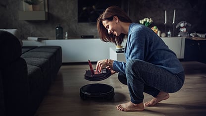 an image of a smiling woman inspecting a small domestic portable device which appears to be an autonomous vacuum cleaner