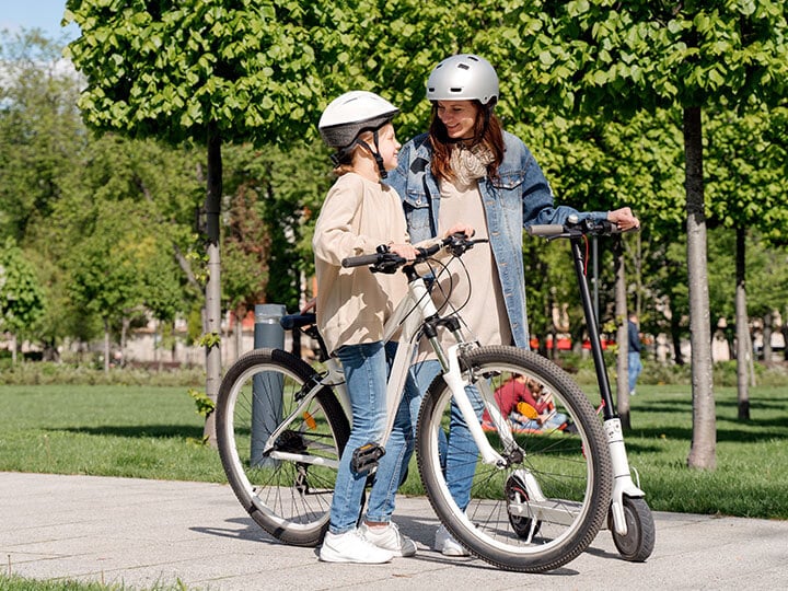 eBike Testing and Certification with mother and daughter looking at each other wearing helmets and standing in a park with an e-bike and e-scooter