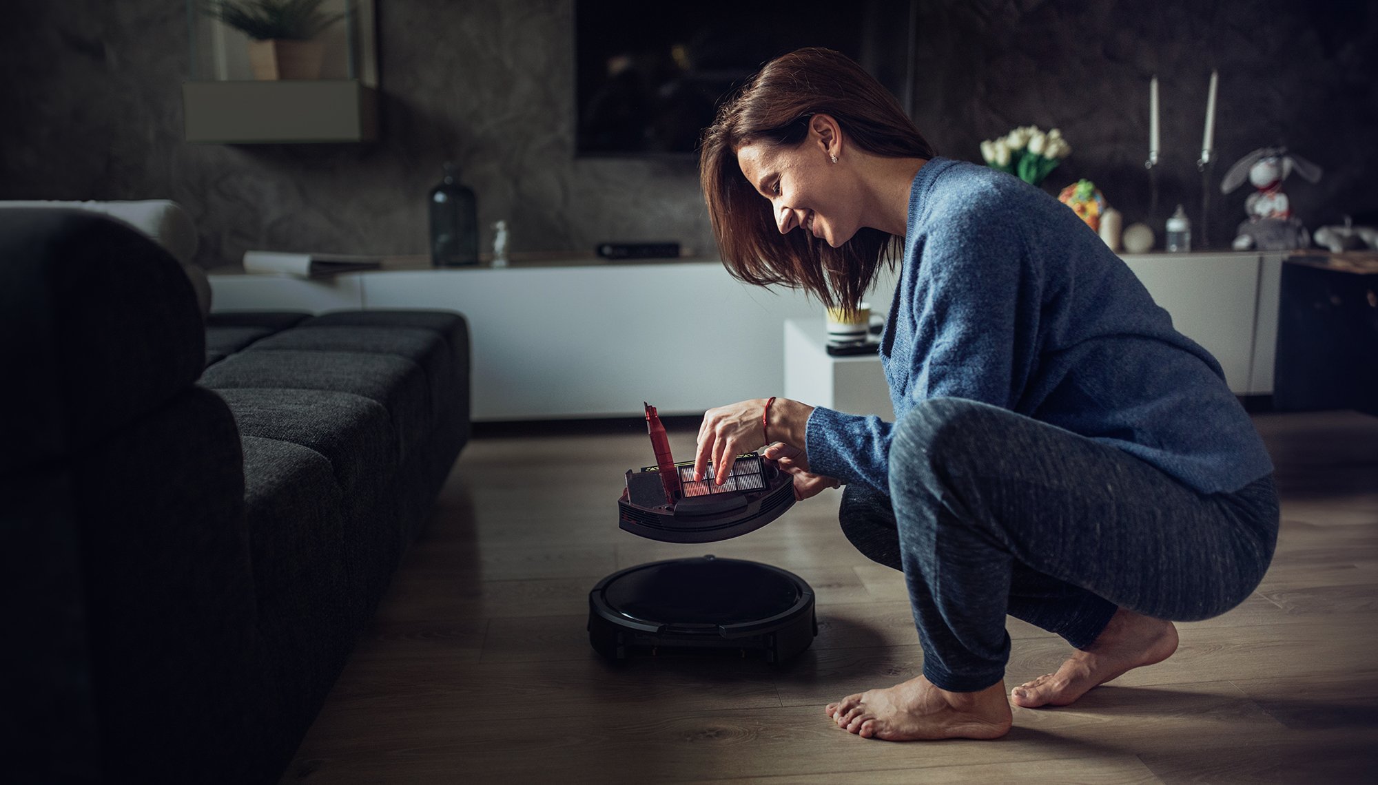 an image of a smiling woman in a domestic situation inspecting batteries in a small device which appears to be a robot vacuum cleaner