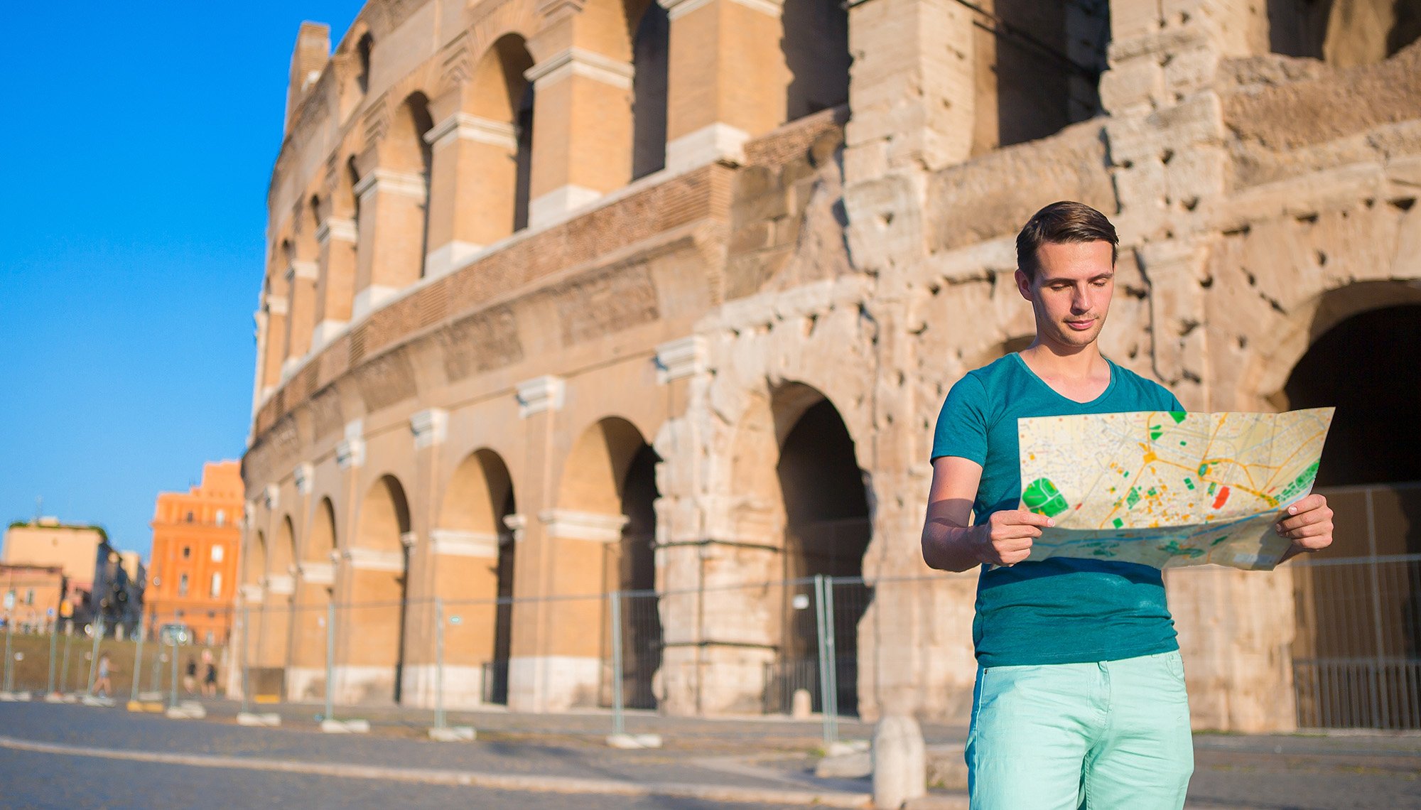 Young man in a teal shirt holding a map while standing in front of the Colosseum