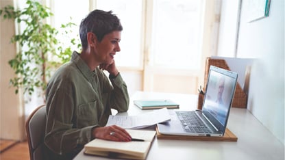 A woman in a green top sits at a laptop in a bright room.