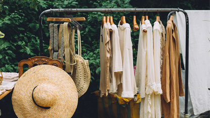Clothes rack with tops, belts, hats, and bags outdoors with trees and table in the background
