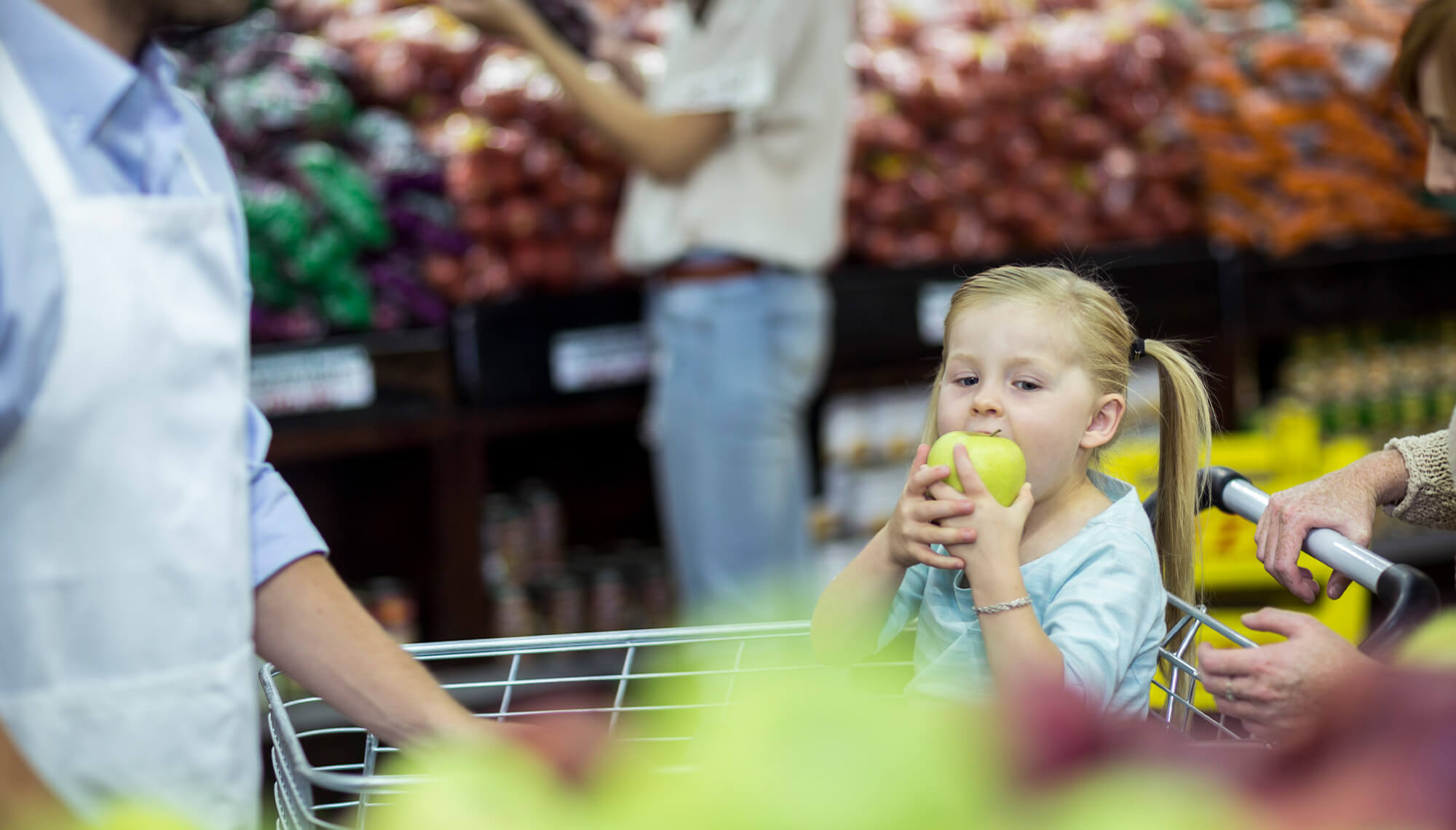 Young girl with blonde pigtails sitting in a grocery cart eating a large green apple