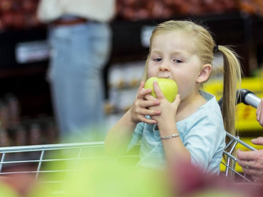 Young girl with blonde pigtails sitting in a grocery cart eating a large green apple