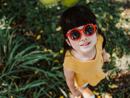 Young girl in a yellow outfit standing in a shaded grove looking up while wearing a pair of large red sunglasses