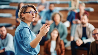 A woman in glasses and a blue top gesticulates as she addresses a group of people in the background.