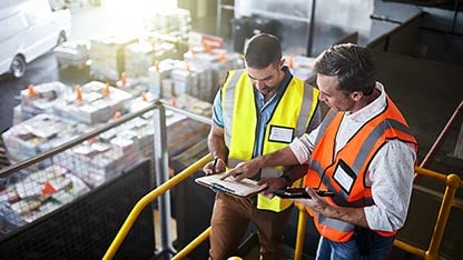 Two men in safety vests, yellow and orange, discussing data points in a notebook while standing in a warehouse 