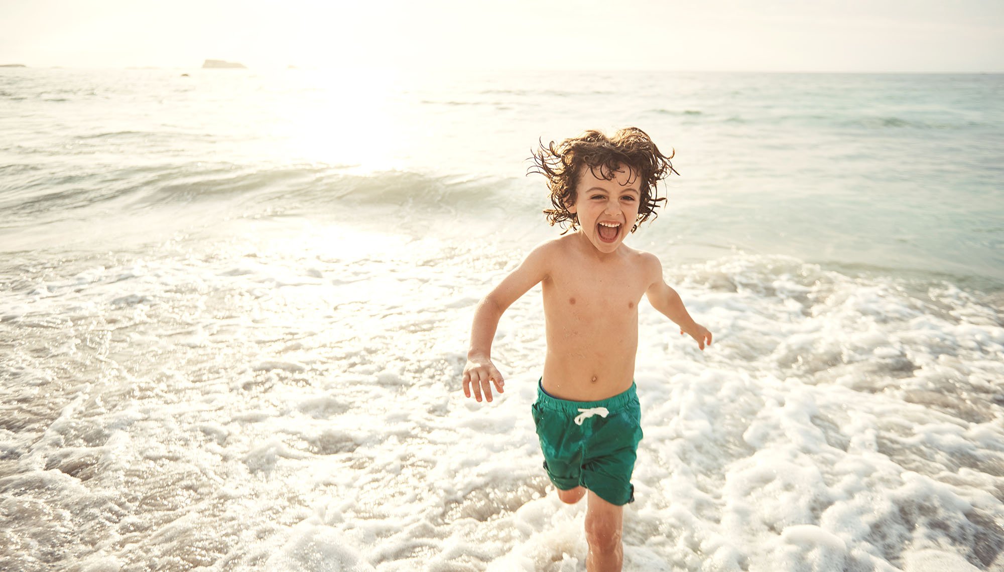 A young boy in green swim trunks laughs and runs through foaming waves on a beach