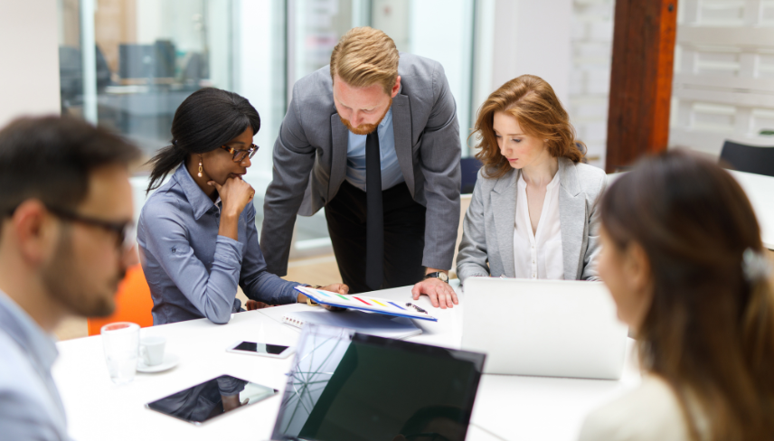 A group of professionals gathered in a meeting room, focused on a laptop screen during a discussion.